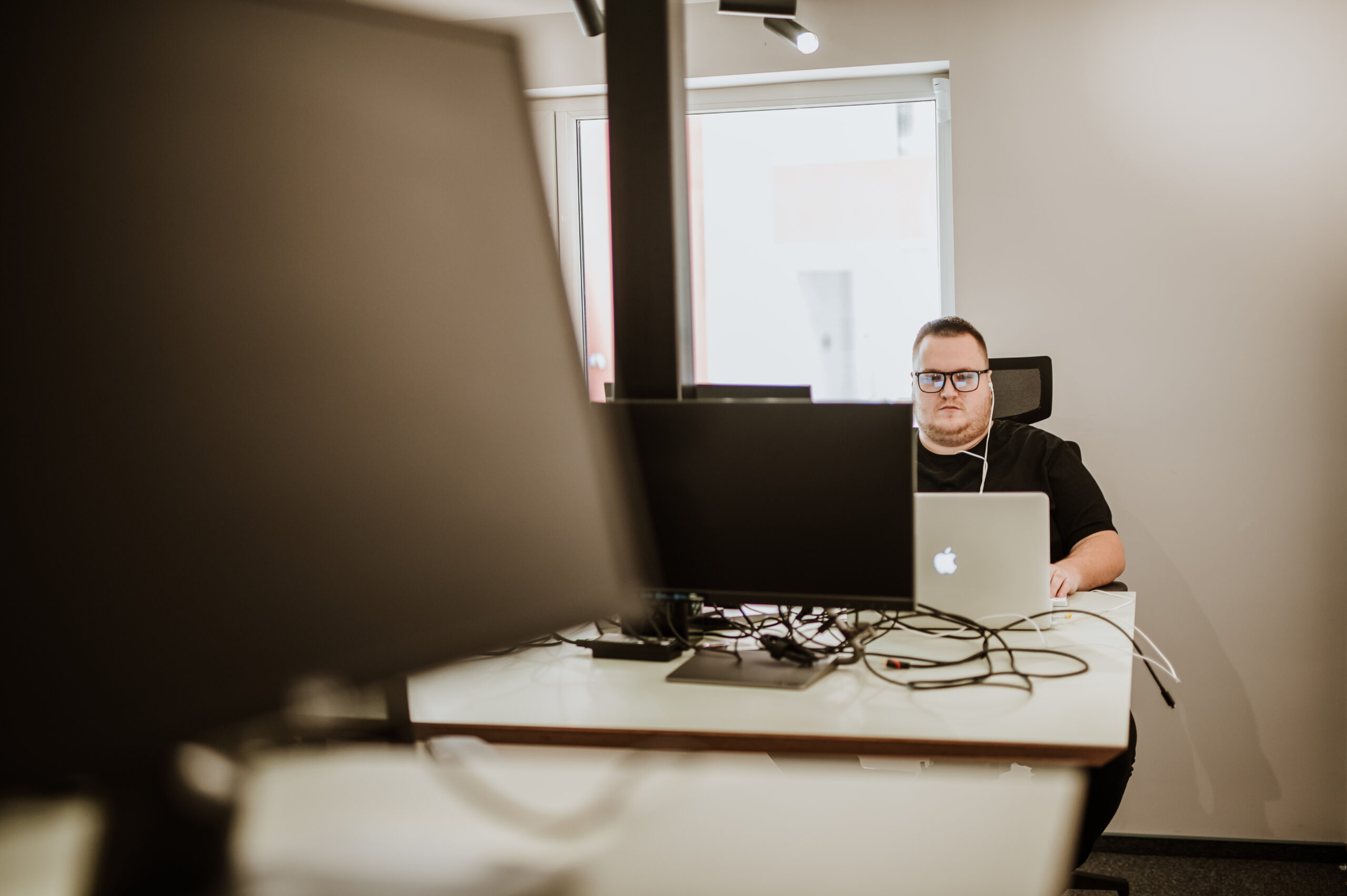 A BetterQA engineer at a laptop behind a row of monitors and cables, seen across the office