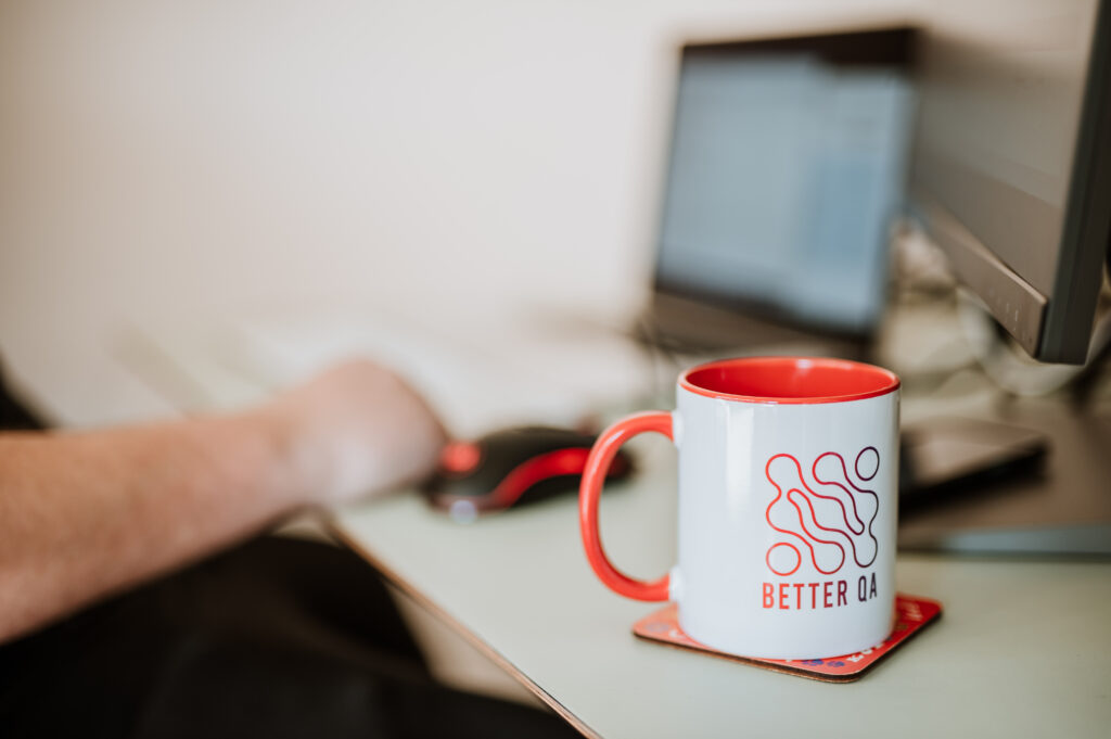 Why feature velocity doesn't matter without independent QA A red BetterQA mug beside a keyboard, a hand on the mouse and a monitor behind