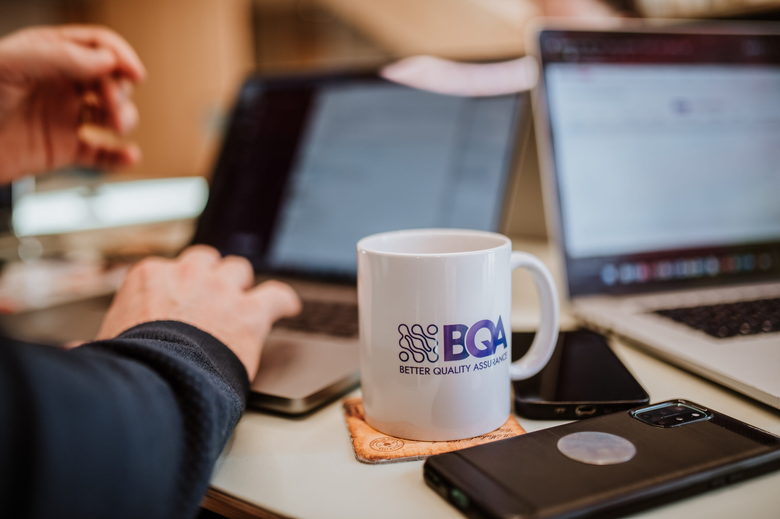 A BetterQA mug beside a colleague's hands working across two laptops