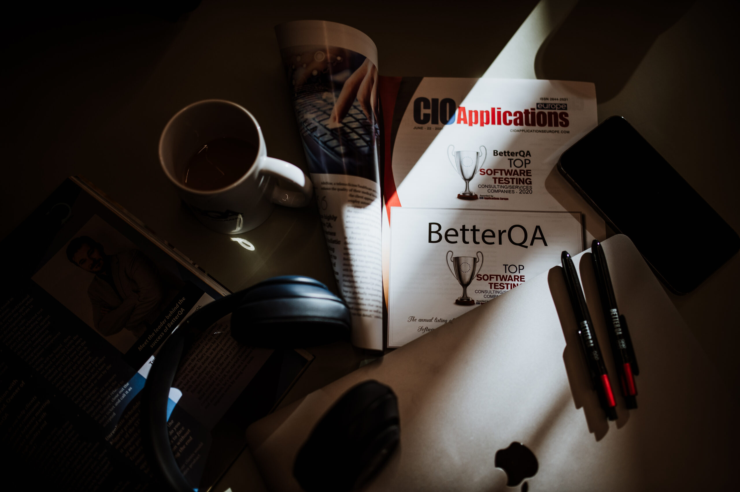 Desk in low light with headphones, a coffee cup and a magazine naming BetterQA a top software testing company