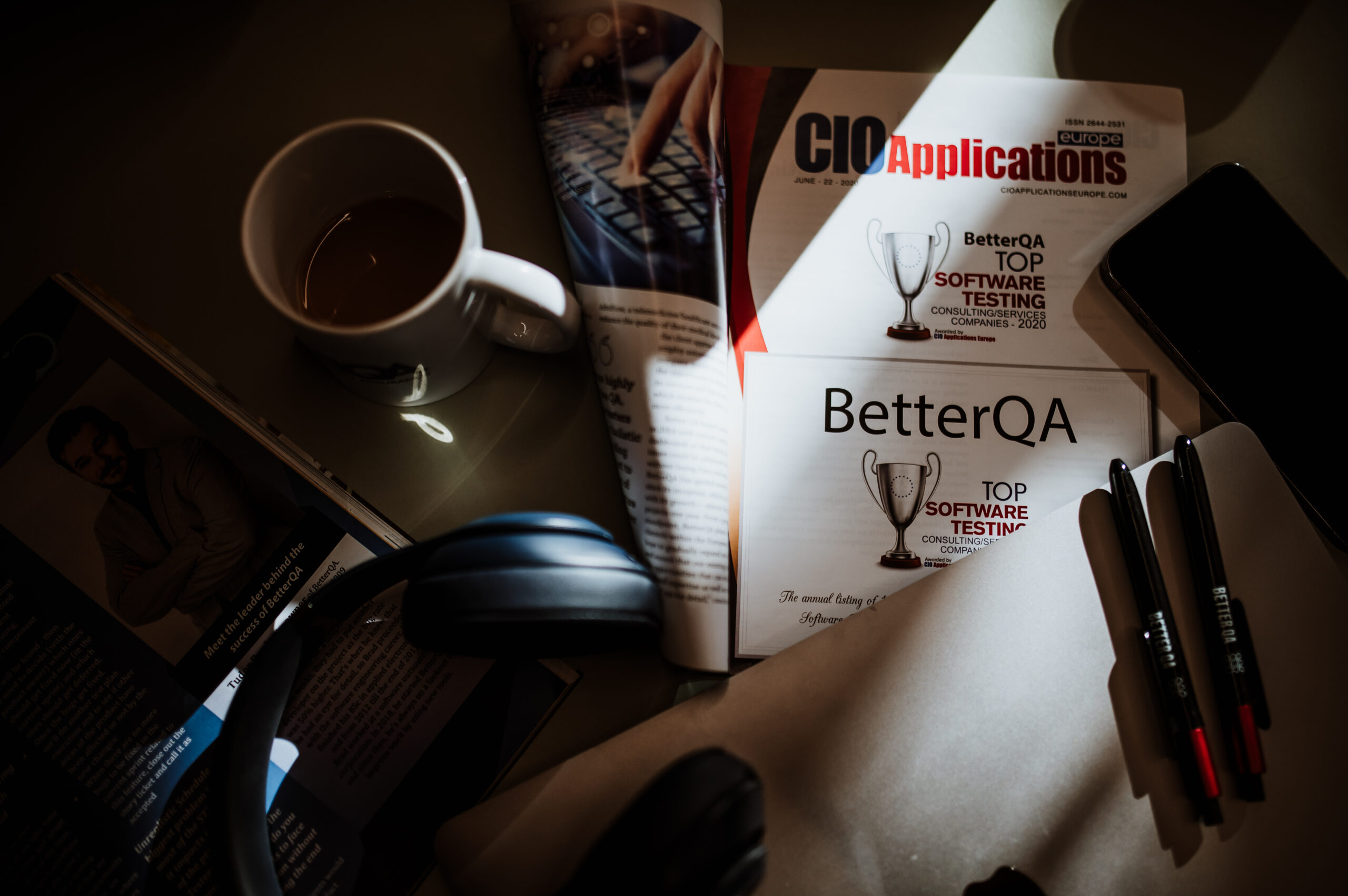 Pens, headphones and a coffee next to a magazine feature naming BetterQA a top software testing company