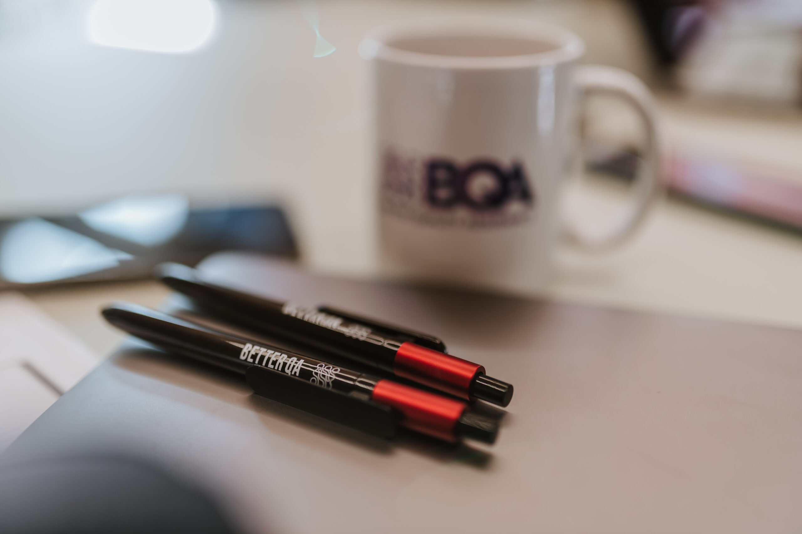A BetterQA mug and branded BetterQA pens on a desk