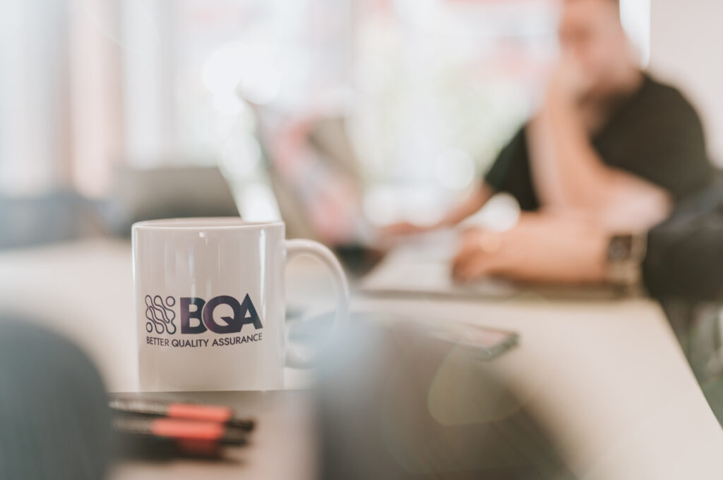 Why testing owned by development is not independent QA A BetterQA mug on a meeting table, with a colleague working at a laptop out of focus behind it